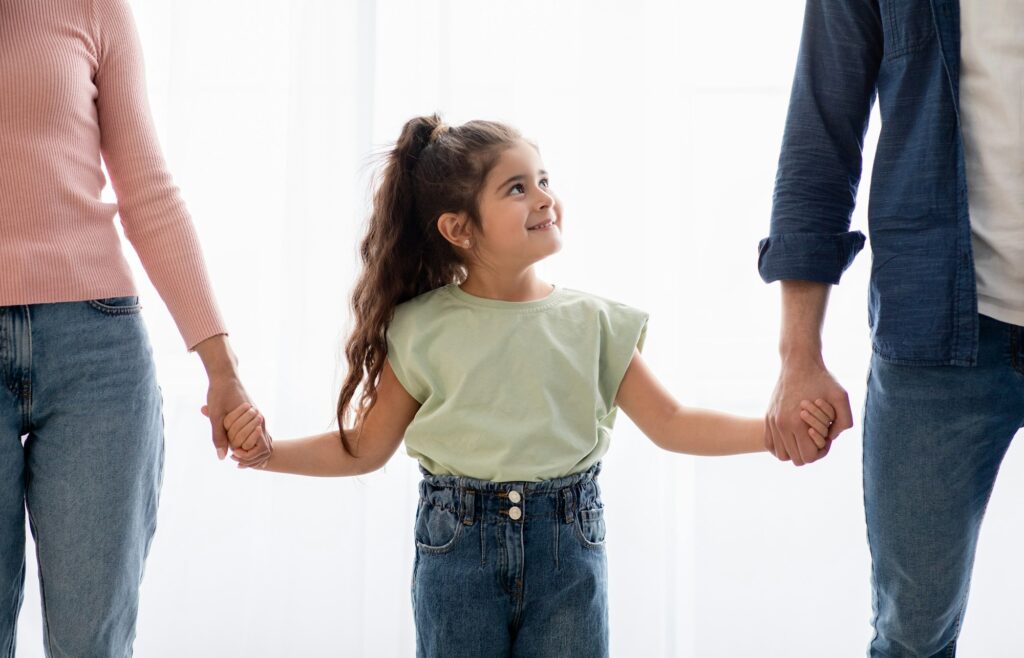 Adoption Concept. Adorable Little Girl Holding Hands With Unrecognizable Mom And Dad