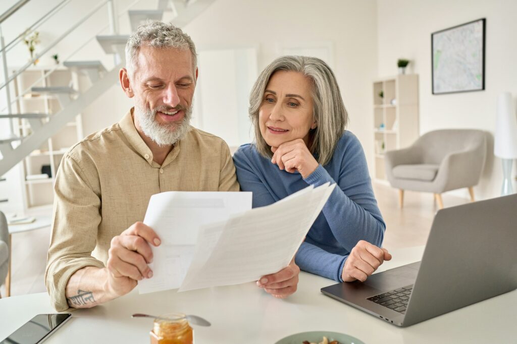 Middle aged couple reading paper bills calculating pension using laptop at home.